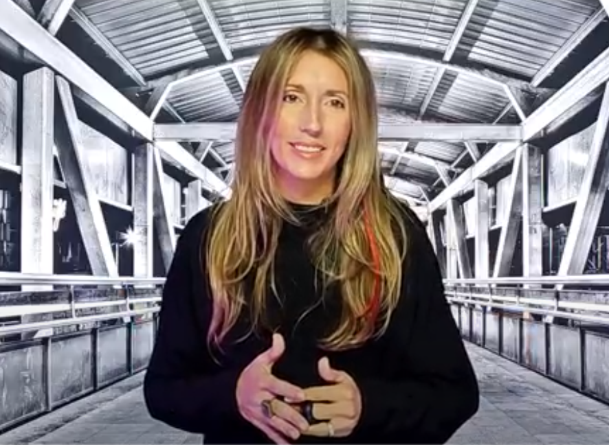 Woman in her late 30's with blond hair, hazel eyes, wearing a black long-sleeved shirt. Image showing woman standing from waste-up with hands in front. Background image makes it look as though the woman is standing in the middle of an old industrial looking bridge and is displayed as black and white.